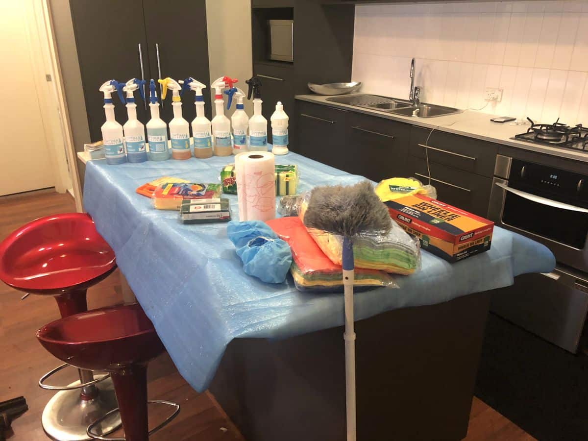 Essential cleaning supplies laid out on a kitchen bench, including spray bottles, cloths, sponges and a duster